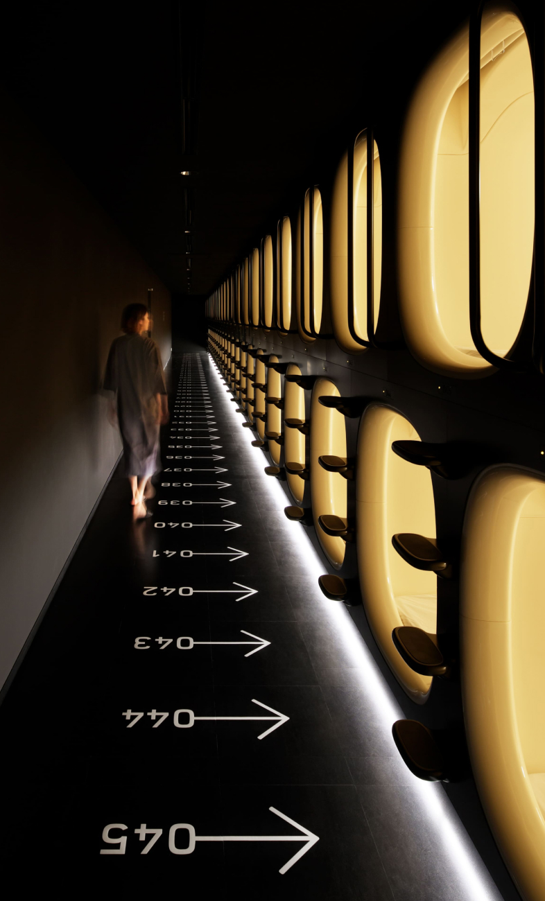 9h nine hours capsule pod hallway at night — a guest walks past illuminated yellow pods stacked floor-to-ceiling